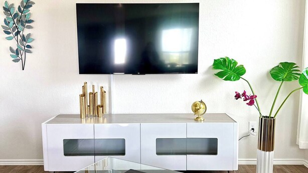 A modern living room with a glass-top coffee table, white entertainment unit, and a flat-screen TV mounted on the wall.