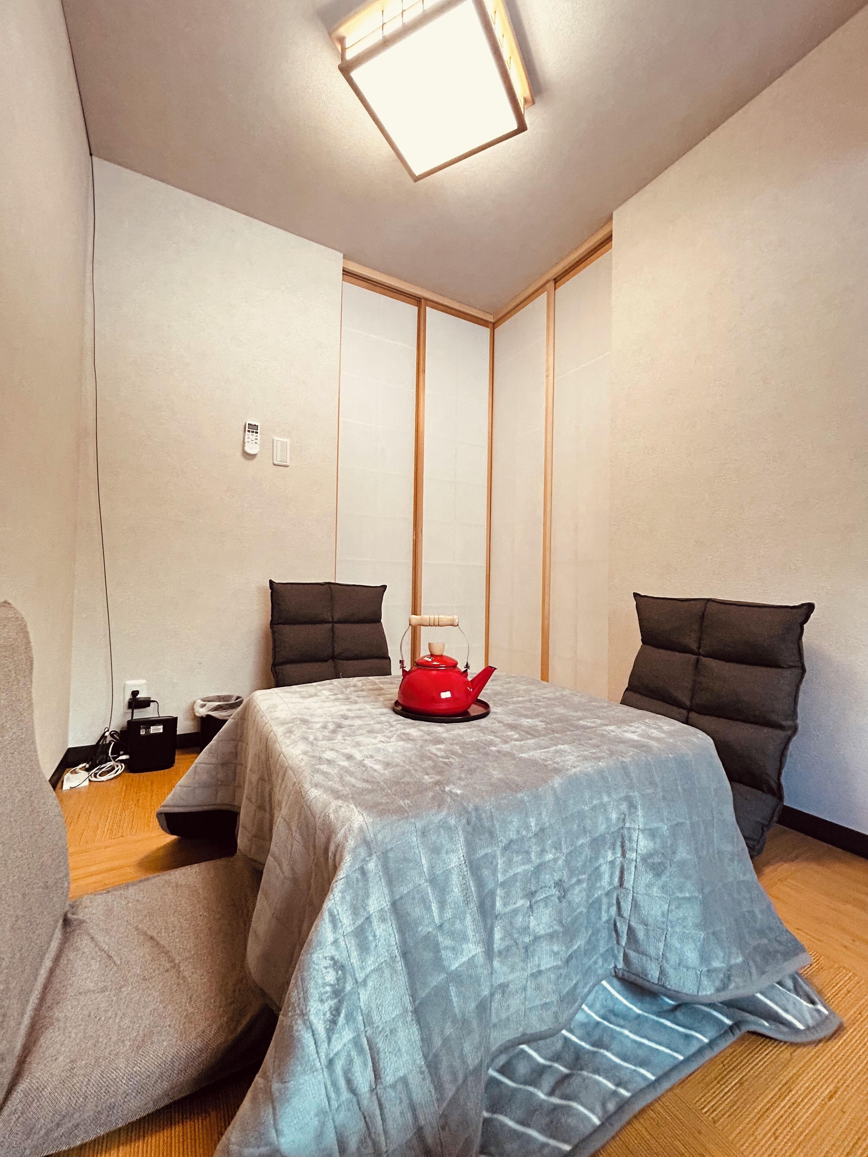 A compact hotel room with a bed, a red kettle, a bedside table, a power outlet, and a wall-mounted light fixture.
