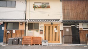 A building with a wooden exterior, a beige facade, and a front door with a sign. There are trash bins and a stack of wooden crates outside. - Miyu Bonichi (Osaka)