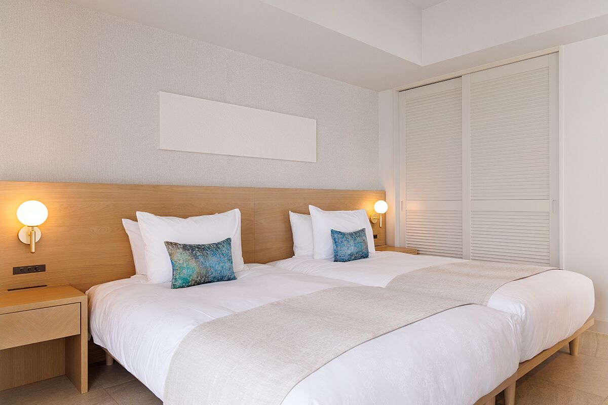 A double bed with white linens and two decorative pillows, a bedside table with a lamp, and a large window with blinds.