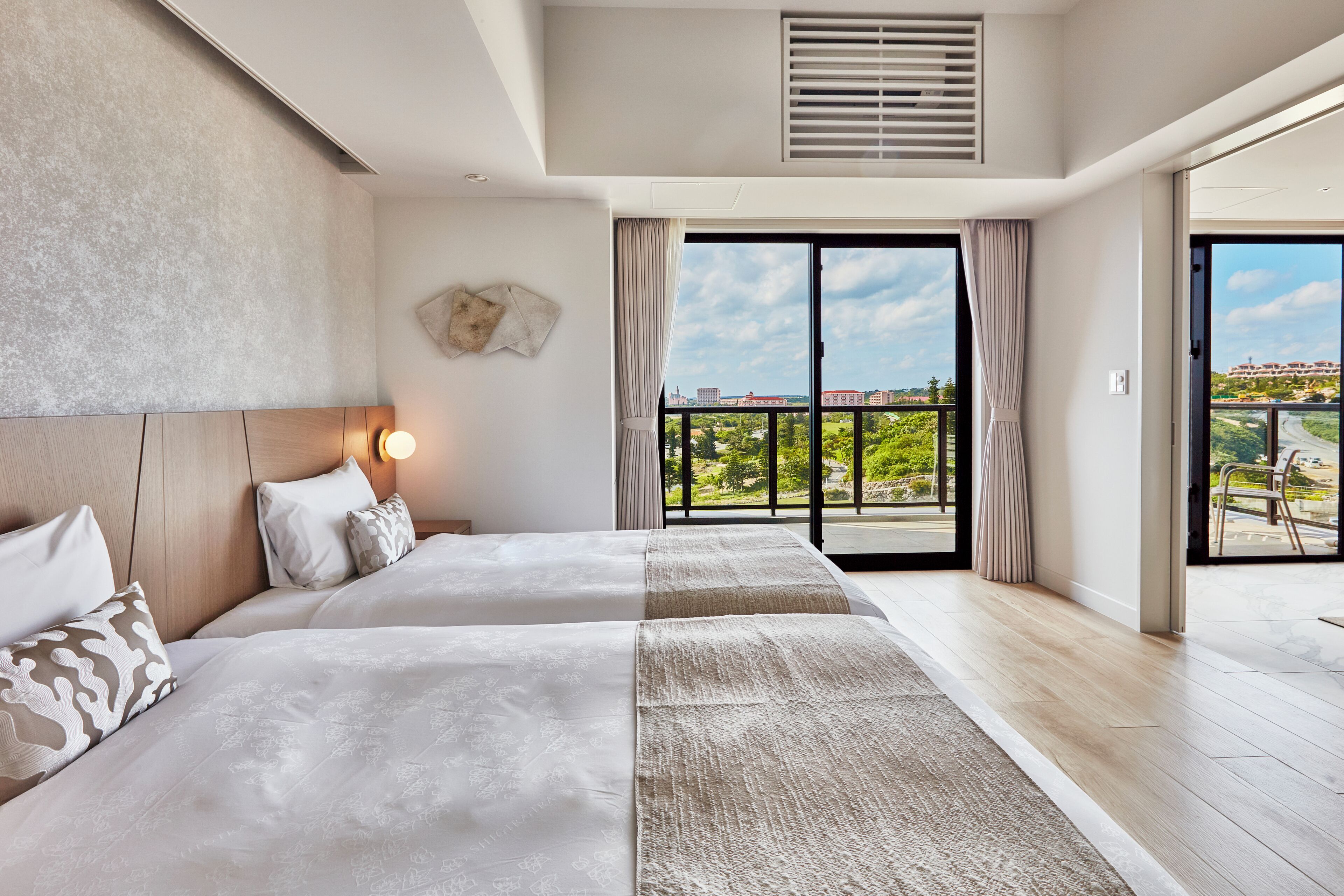 Habitación de hotel moderna con una cama grande, una mesita de noche con lámpara, y vistas a zonas verdes y edificios a través de las puertas del balcón.