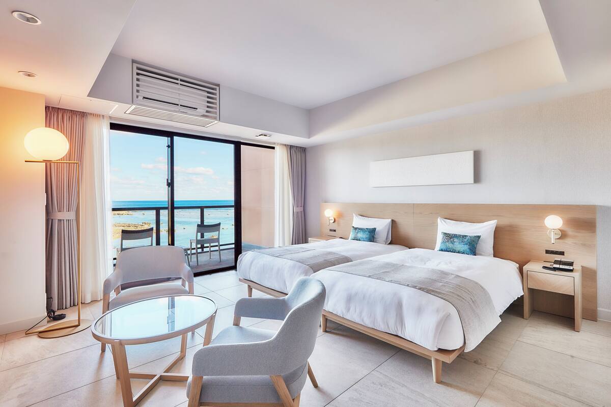 Une chambre d’hôtel avec deux lits, un balcon donnant sur la mer, une petite table et deux chaises.