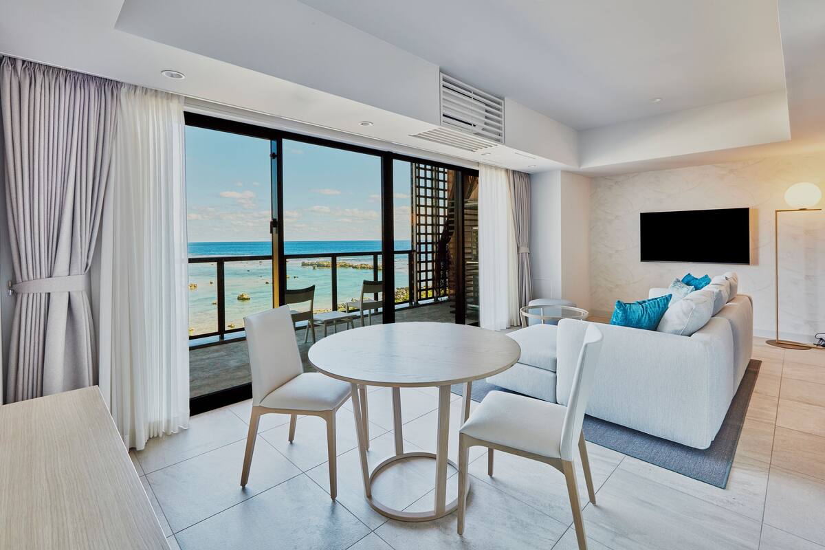 Une chambre d’hôtel moderne avec un balcon donnant sur l’océan, une table à manger ronde avec des chaises, un canapé blanc et un téléviseur à écran plat fixé au mur.
