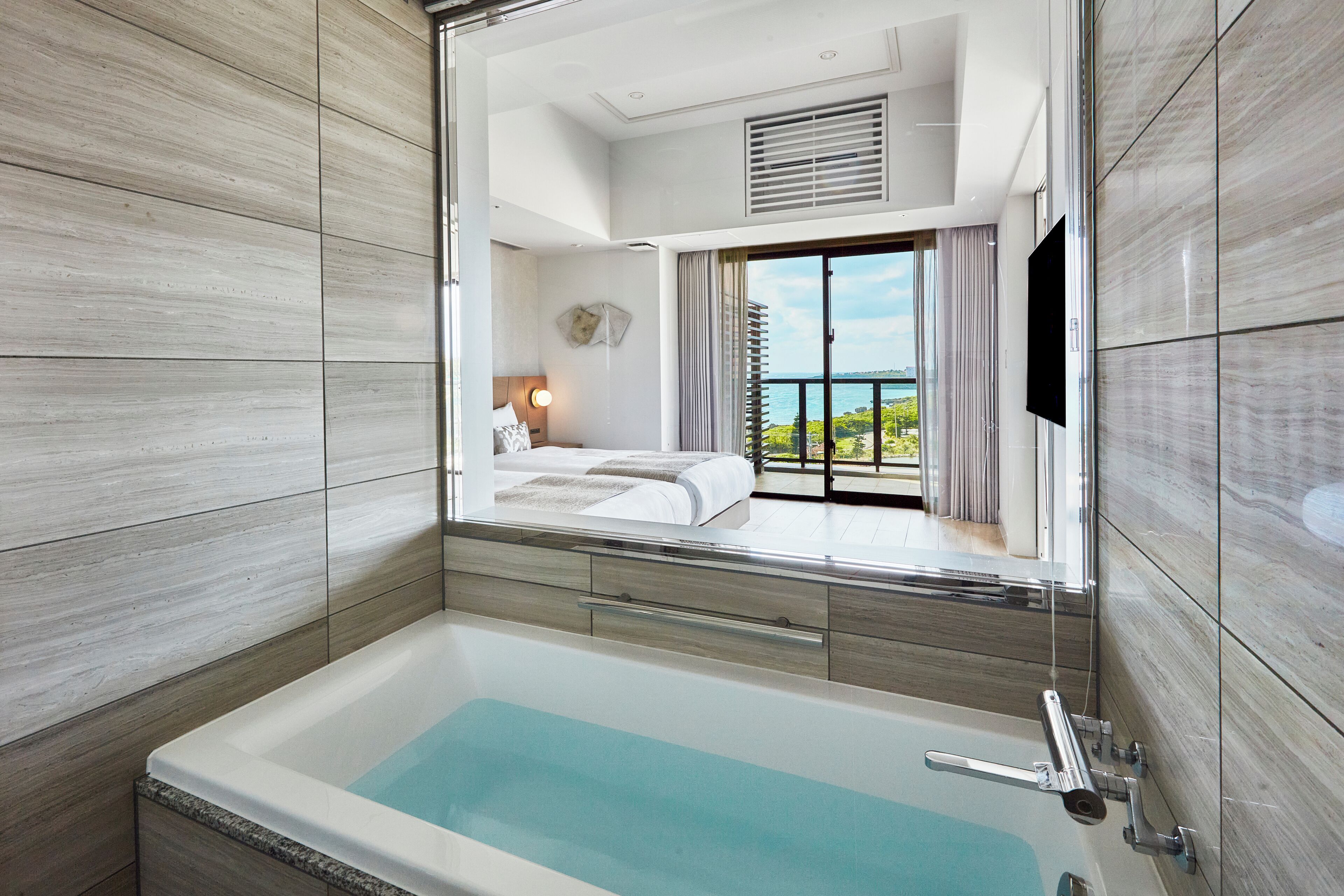 A modern bathroom with a large bathtub, a visible bedroom with a balcony, and a TV mounted on the wall.