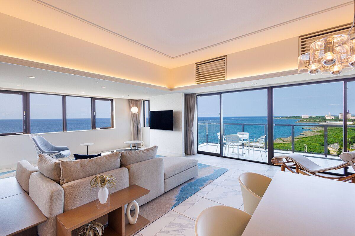Un salon moderne avec un canapé, une table basse et un téléviseur. La pièce est dotée de grandes fenêtres offrant une vue sur la mer et d’un balcon avec des chaises.