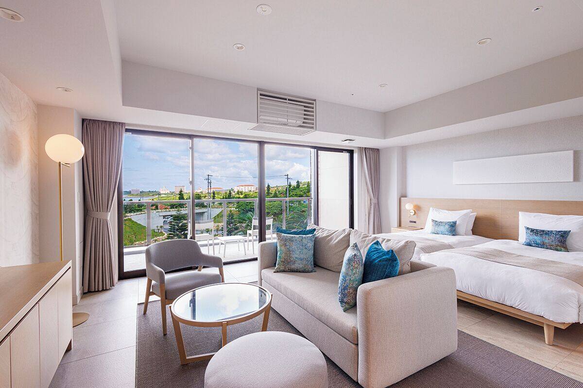 Une chambre d’hôtel moderne, dotée d’un grand lit, d’un canapé, d’une petite table et d’une chaise. Une grande fenêtre offre une vue sur la ville.