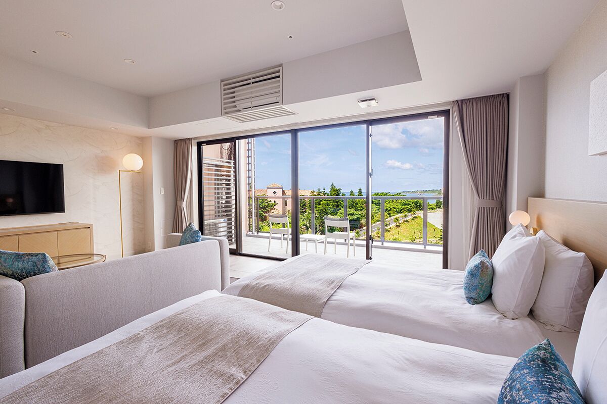 Ein Hotelzimmer mit zwei Betten, einem Flachbildfernseher, einem Schreibtisch und einem großen Fenster mit Blick ins Freie.