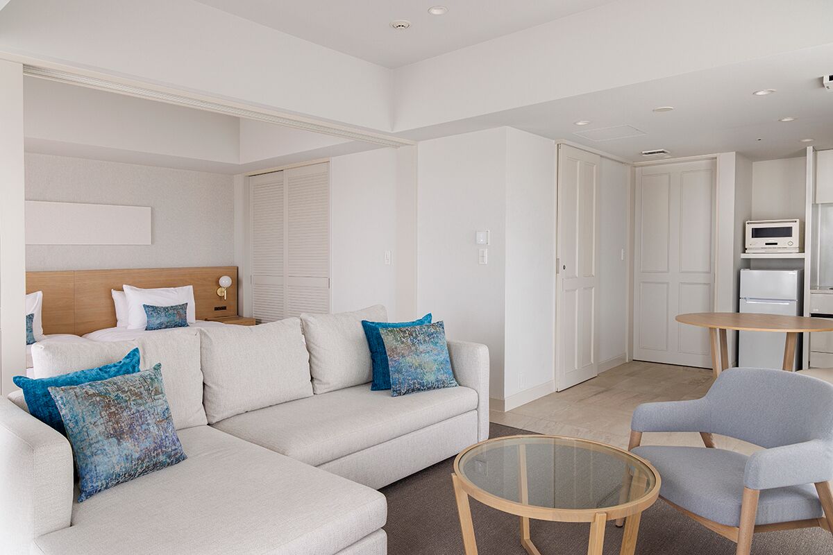 A modern hotel room with a sofa, armchairs, a glass coffee table, and a kitchenette.