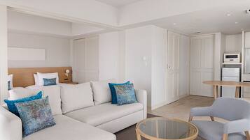 A modern hotel room with a sofa, armchairs, a glass coffee table, and a kitchenette.