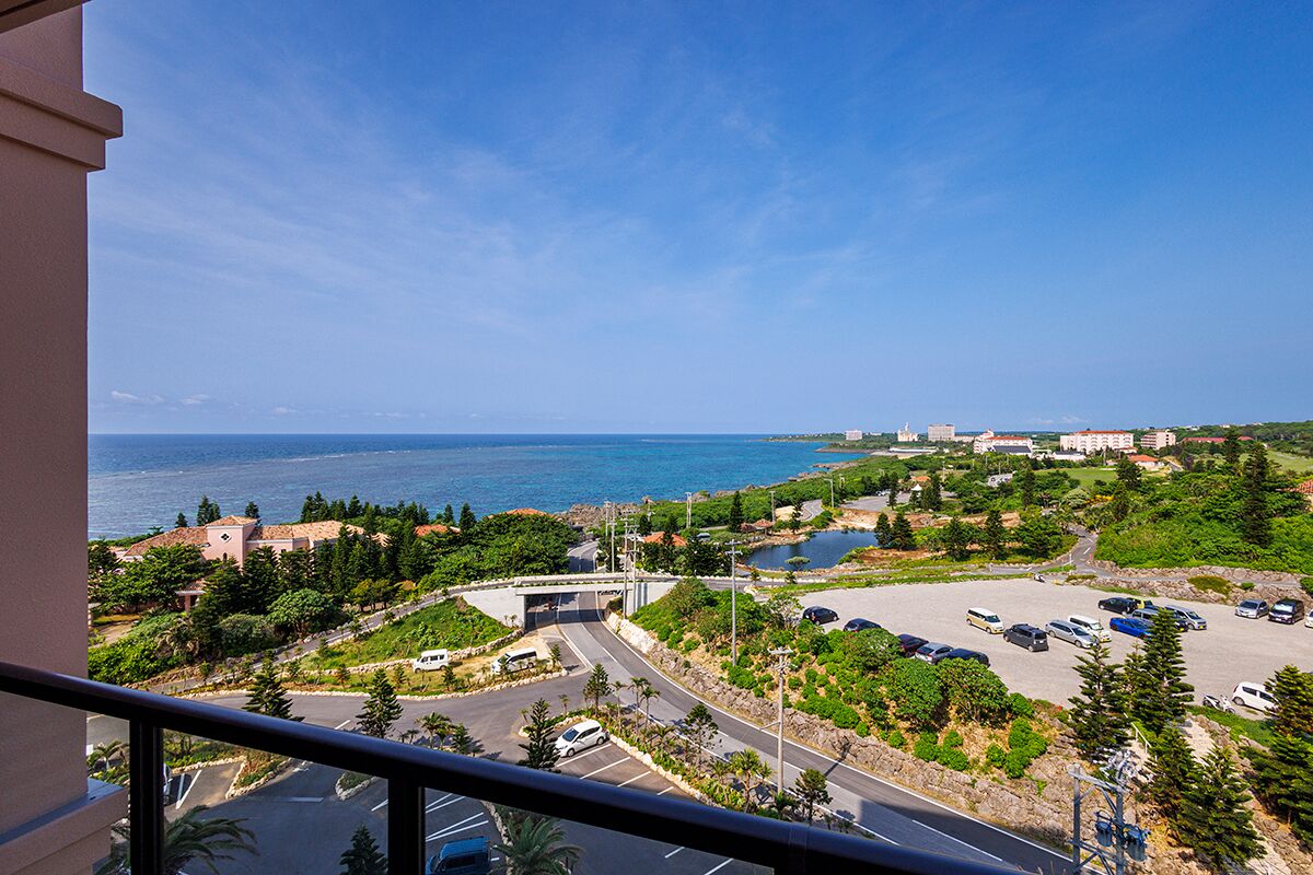A coastal view from a balcony overlooking a parking lot, a road, and a bridge leading to a beach.
