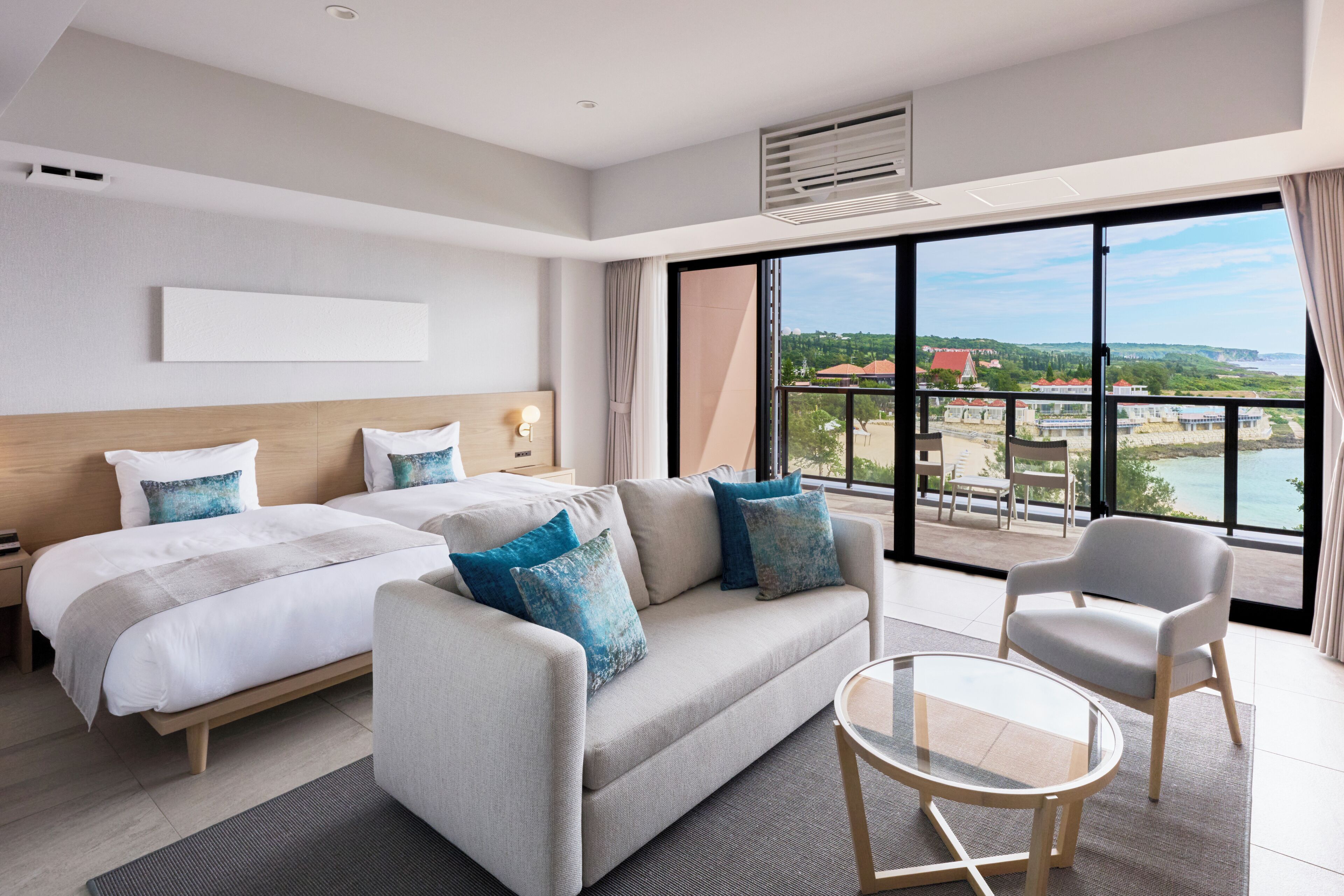 A hotel room with two beds, a sofa, a chair, a glass table, and a large window with a view of a cityscape.