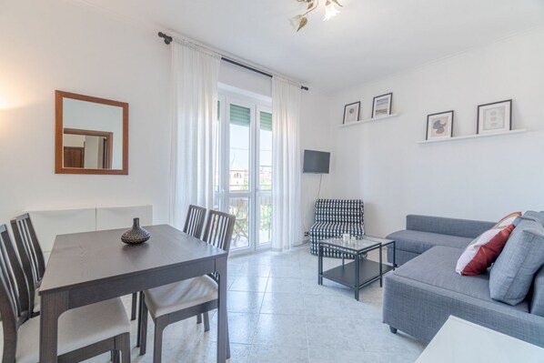 Een moderne woonkamer met een eettafel, stoelen, een bank, een salontafel, een televisie en een balkon met gordijnen.