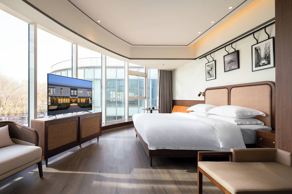 Una habitación de hotel moderna con una cama grande, un sofá, un escritorio y un televisor mostrando un paisaje urbano.
