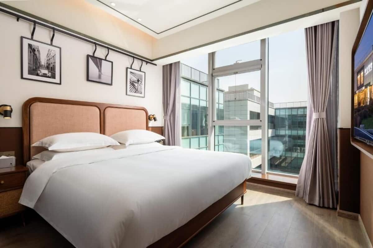 Una habitación de hotel moderna con una cama grande, una mesita de noche, un televisor montado en la pared y un ventanal con vistas a la ciudad.