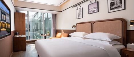 Una camera d'hotel moderna con un letto grande, un comodino, una lampada da tavolo, una finestra con vista su un edificio e quadri appesi alle pareti.
