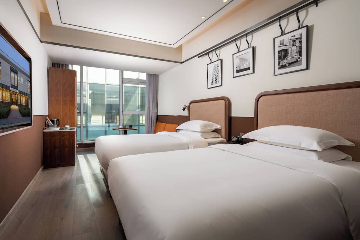 Habitación de hotel con dos camas, una mesita de noche de madera, una pequeña mesa con una lámpara y cuadros enmarcados en la pared.