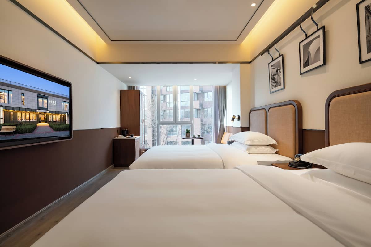 Habitación de hotel con dos camas, un televisor montado en la pared, una ventana con vistas a edificios y una mesita con una lámpara.