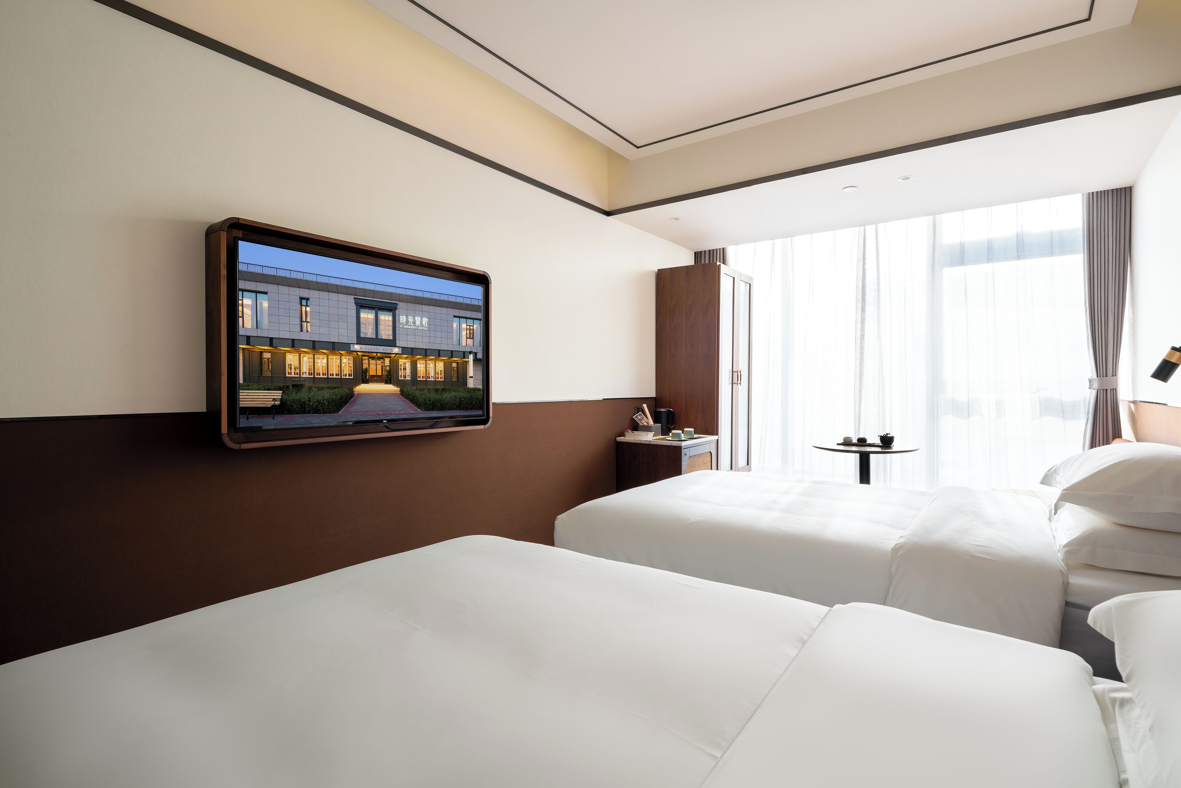 Ein Hotelzimmer mit zwei Betten, einem an der Wand montierten Fernseher, einem kleinen Tisch mit Lampe und einem Fenster mit Vorhängen.