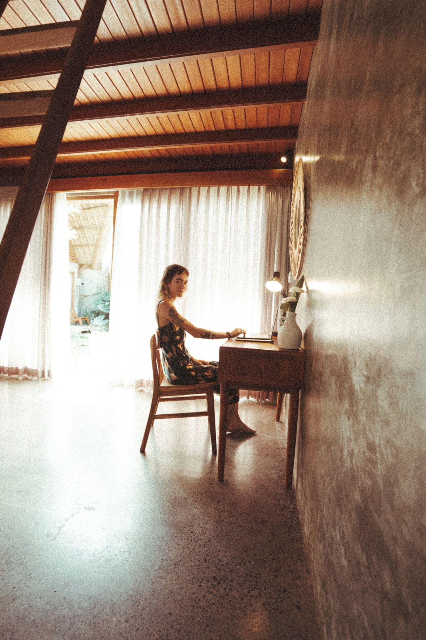 A room with a wooden ceiling, a desk with a lamp, and a chair.