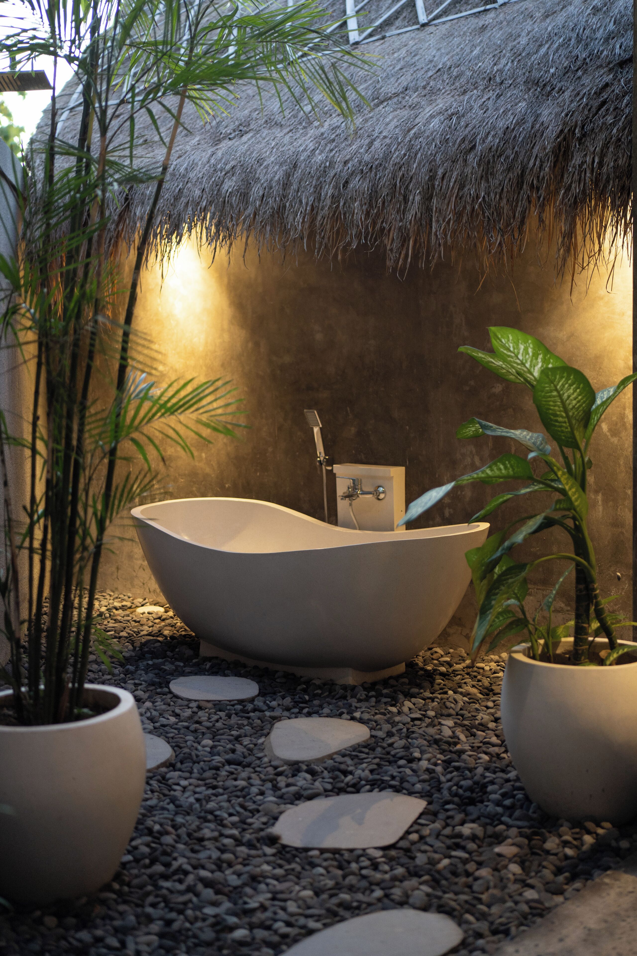Un baño moderno con una bañera independiente, rodeado de piedras y plantas en macetas, y una estructura de techo de paja al fondo.