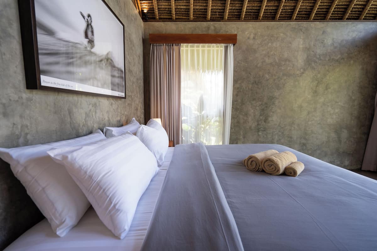 A neatly made bed with white linens and rolled towels, a framed picture on the wall, and a window with curtains.