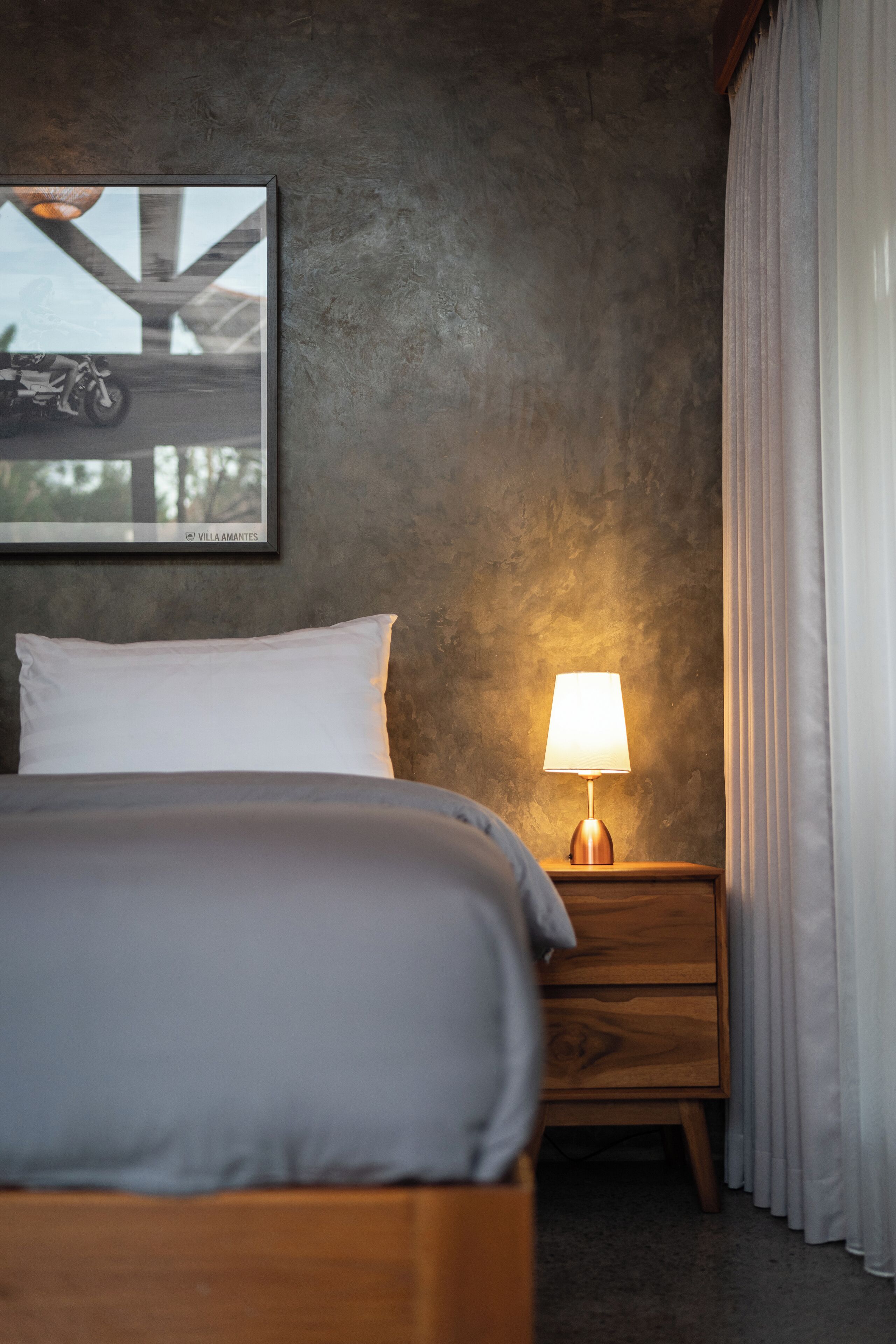 Ein ordentlich bezogenes Bett mit weißer Bettwäsche, ein Nachttisch aus Holz mit einer Lampe und ein gerahmtes Bild an der Wand.