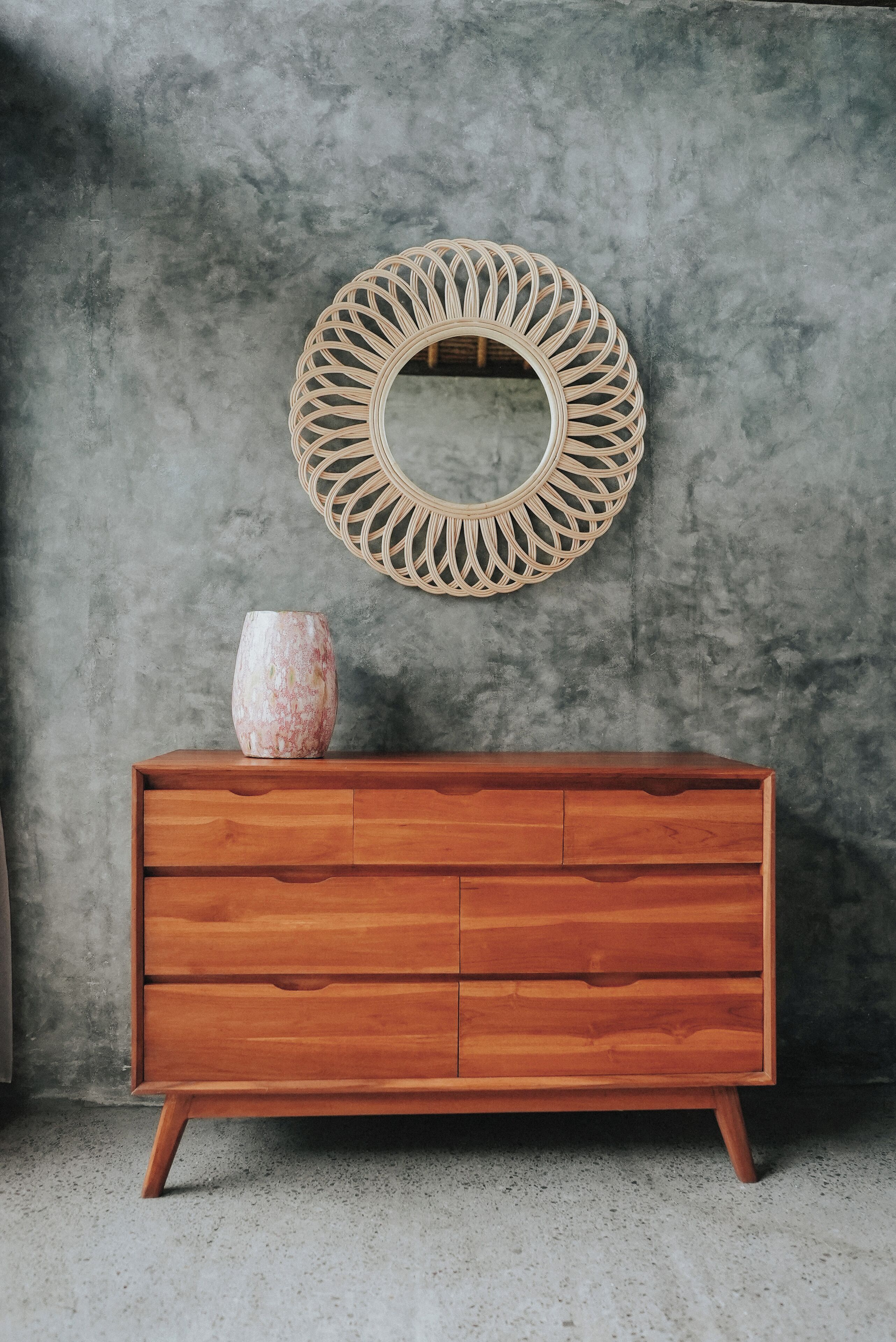 Un aparador de madera con cajones y un florero encima, frente a una pared con textura y un espejo circular arriba.