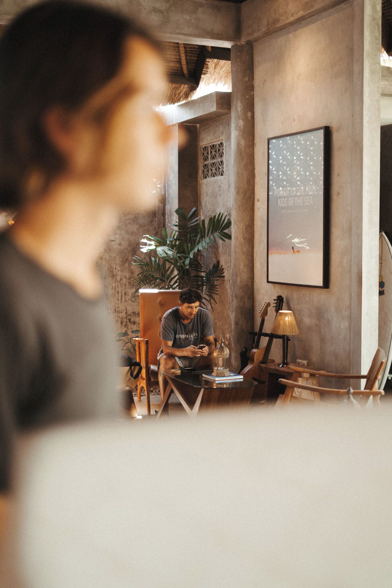 A person sitting at a table with a laptop, surrounded by plants and a guitar, in a room with a concrete wall and a framed picture.