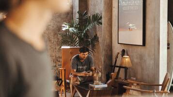 A person sitting at a table with a laptop, surrounded by plants and a guitar, in a room with a concrete wall and a framed picture.
