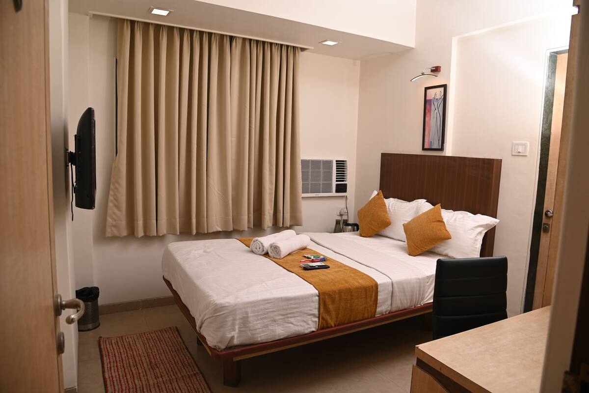 A hotel room with a bed, a desk, a chair, a TV, a window with curtains, an air conditioning unit, and a painting on the wall.