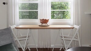 Une pièce avec une table en bois et deux chaises blanches, une petite sculpture en bois posée sur la table, et une vue sur la verdure à travers de grandes fenêtres.