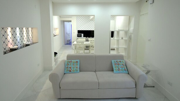 Ein modernes Wohnzimmer mit einem weißen Sofa und bunten Zierkissen, im Hintergrund eine Küche und ein Essbereich mit weißer Möbel.