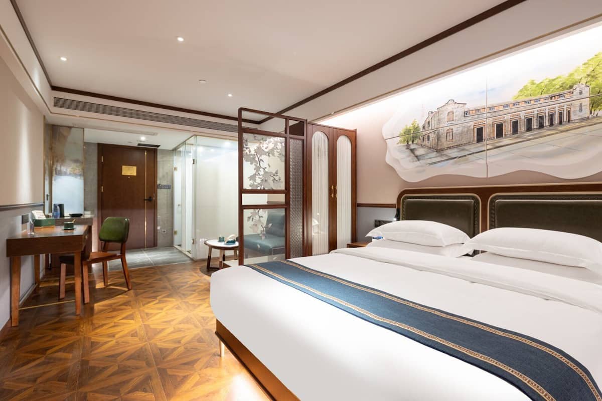 A hotel room with a large bed, a desk, a chair, and a view of a building through a window.