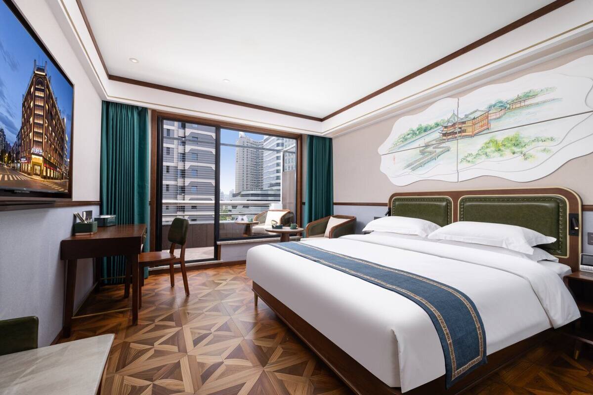 A hotel room with a large bed, a desk, a chair, and a view of a cityscape through a large window.