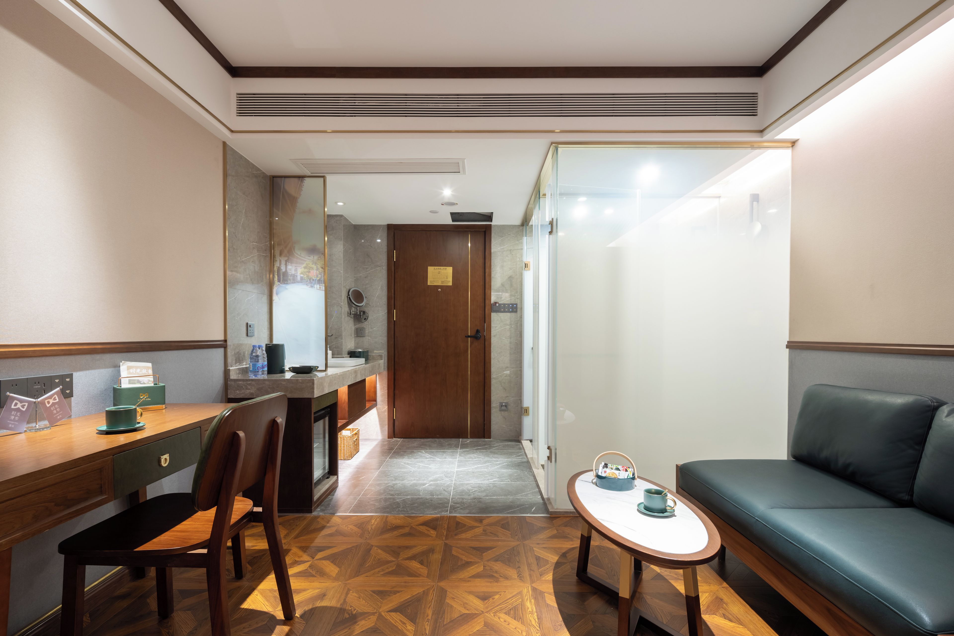 A hotel room with a wooden desk, a chair, a small table with a cup, a sofa, and a door leading to a bathroom.