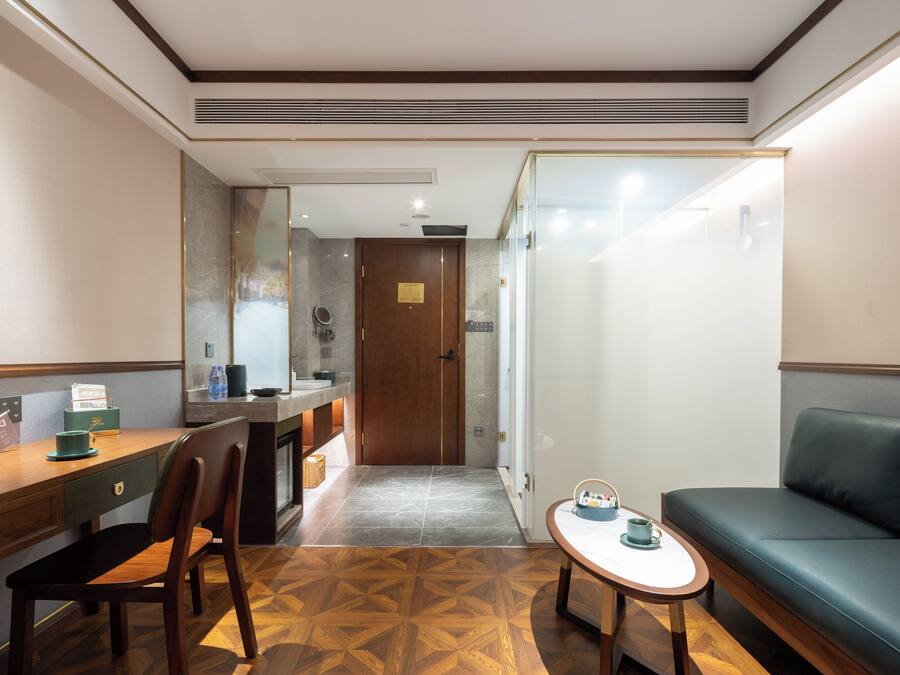 A hotel room with a wooden desk, a chair, a small table with a cup, a sofa, and a door leading to a bathroom.