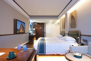 Une chambre d’hôtel avec un grand lit, un bureau avec une tasse et un panneau, une chaise, une petite table avec un panier, et un mur orné d’un tableau encadré.