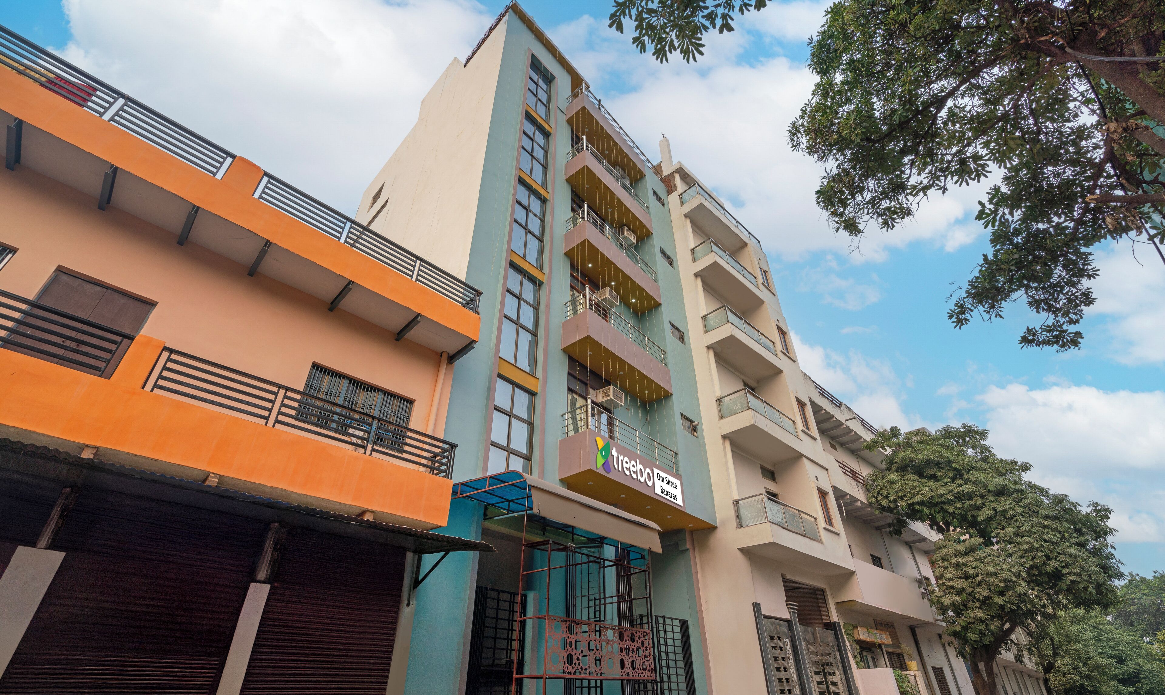 A multi-story building with balconies, a sign for 'Trebo Om Shree Banaras', and a tree in the foreground.