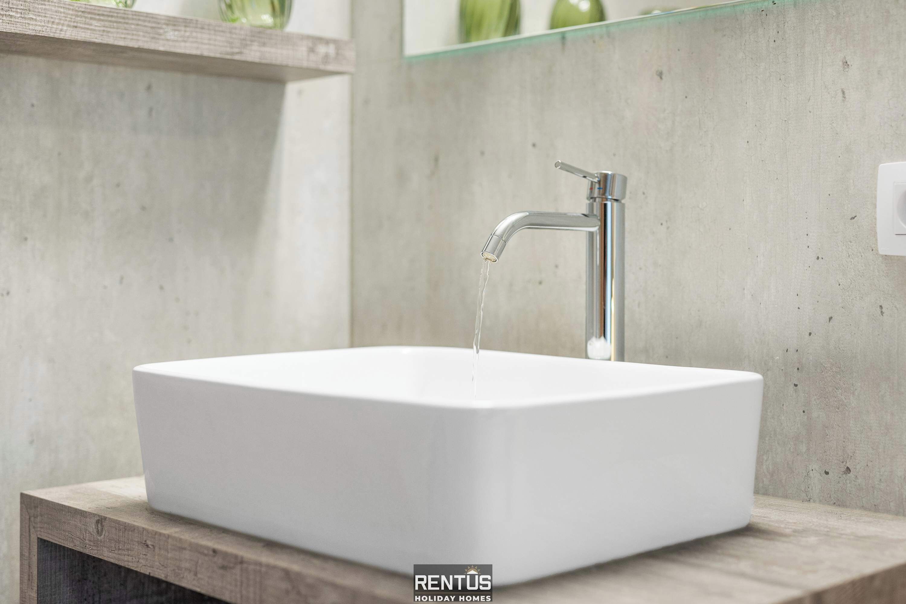Ein modernes Badezimmer mit einem rechteckigen weißen Waschbecken, einem chromfarbenen Wasserhahn und einem Glasregal darüber.