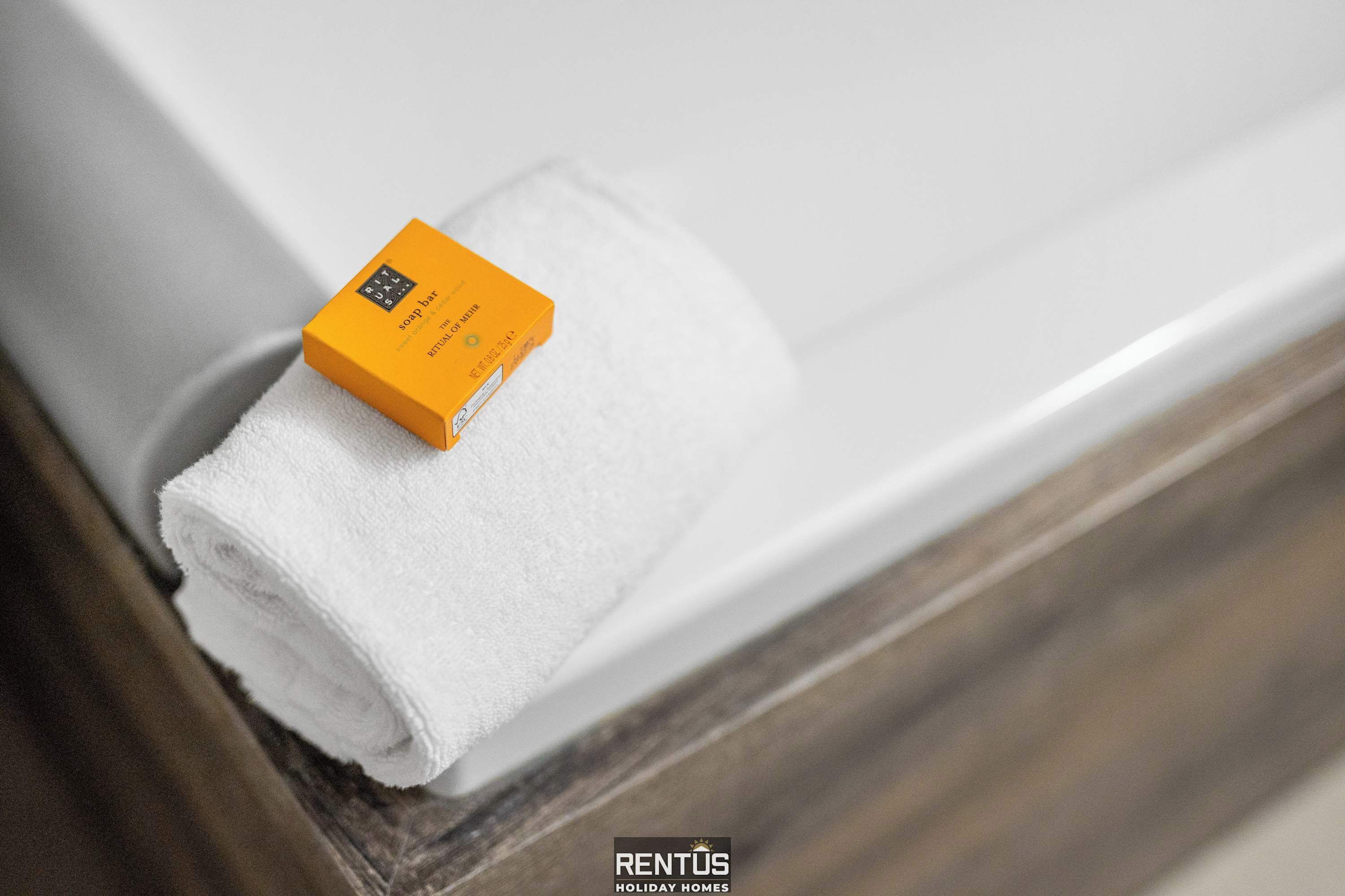 A rolled white towel on a wooden surface, likely a bathroom counter, with a small orange box labeled "soap bar" on top.