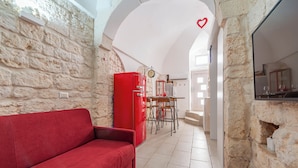 Un intérieur chaleureux avec un canapé rouge, des murs en pierre, un coin repas avec des chaises et un espace cuisine doté d’un réfrigérateur rouge.