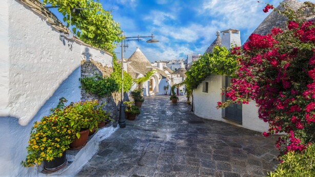Une rue étroite, pavée de pierres, bordée de bâtiments blancs aux portes en arc et à l’architecture traditionnelle.