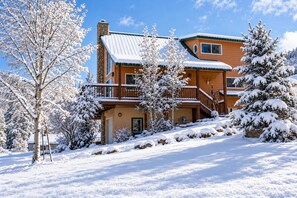 A two-story lodge with a stone chimney, a balcony, and snow-covered trees. - Creekside Cabin: Pet Friendly, Hot tub, Sled Hill, Walk to Shops, Dining & Springs, 2 Living Spaces (Pagosa Springs)