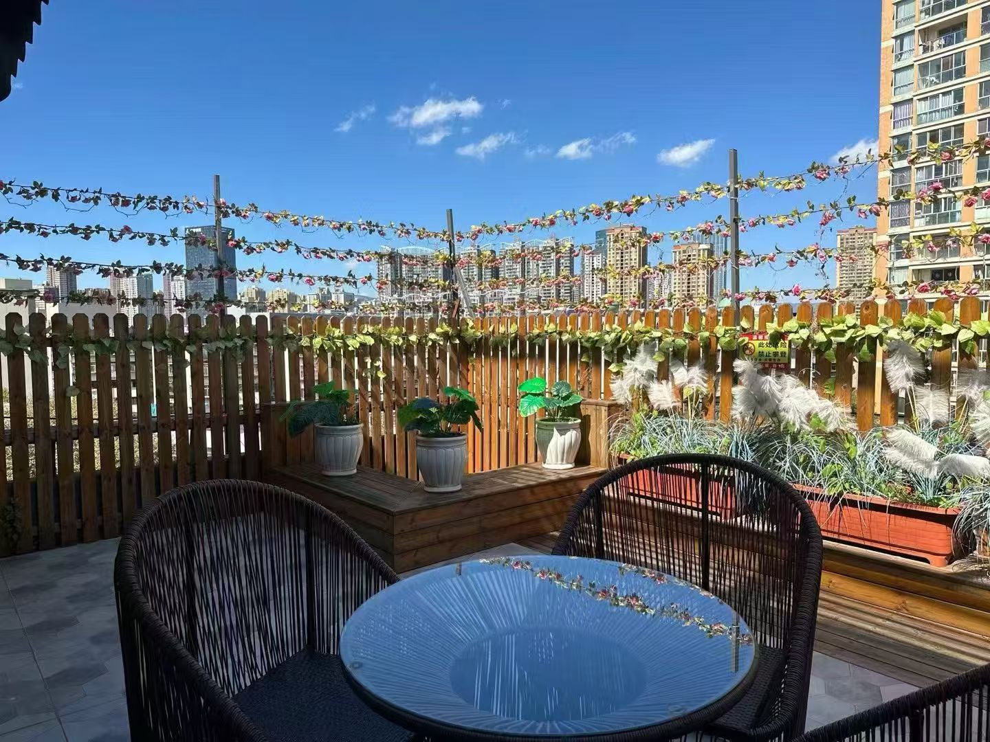 Un patio al aire libre con mobiliario de mimbre, plantas en macetas y vistas a un paisaje urbano.