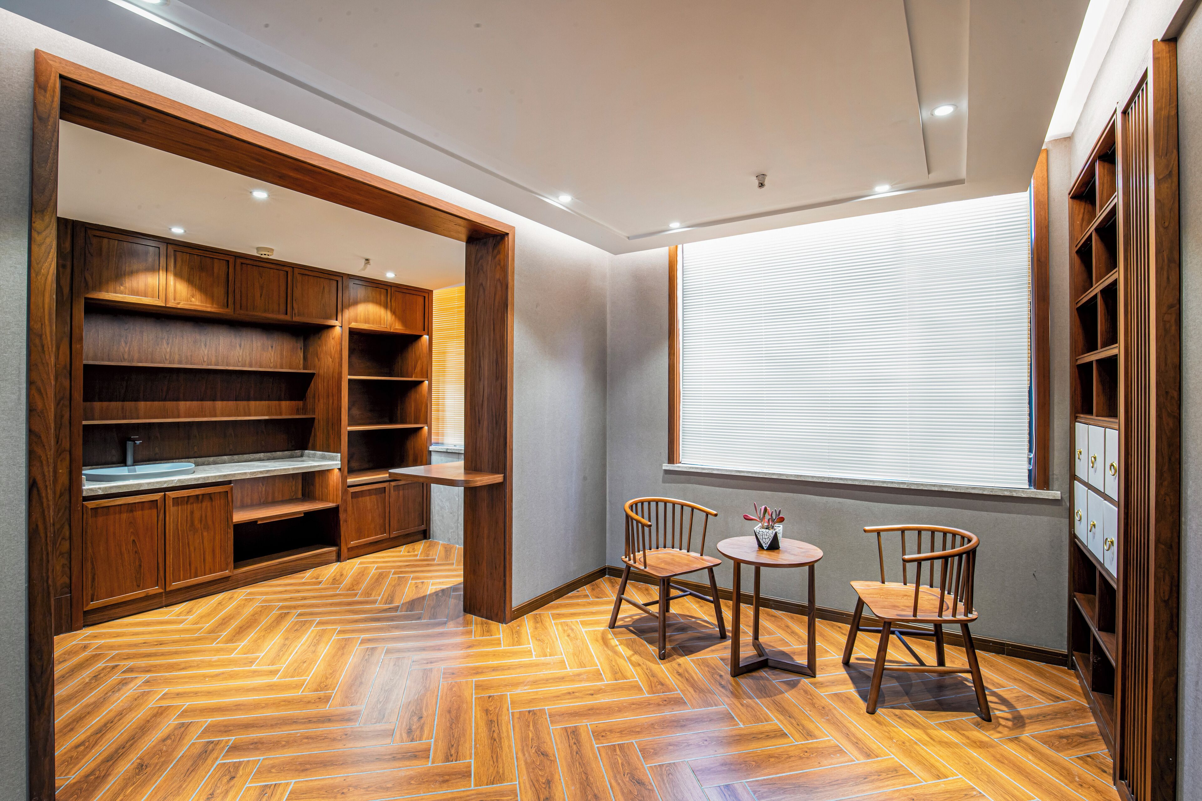 Una habitación con estanterías de madera, suelo de espiga, dos sillas, una mesita y una ventana con persianas.