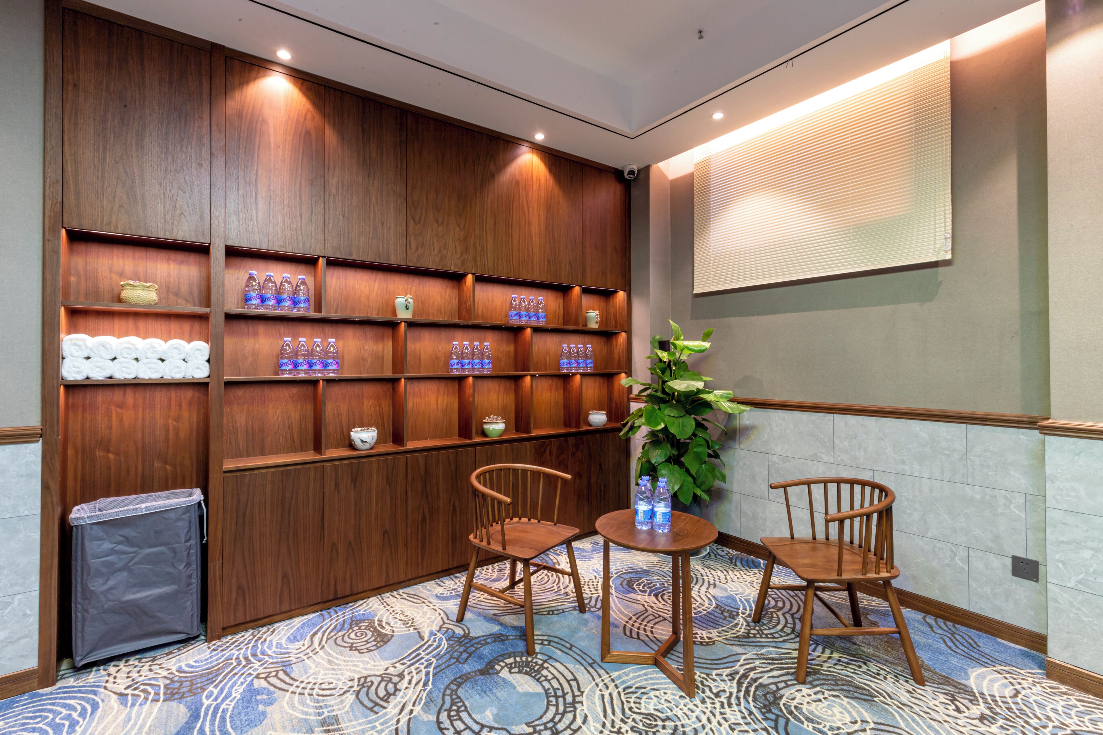 Et hotel lobby med træpaneler, et mønstret tæppe, to træstole, et lille bord og en skraldespand.