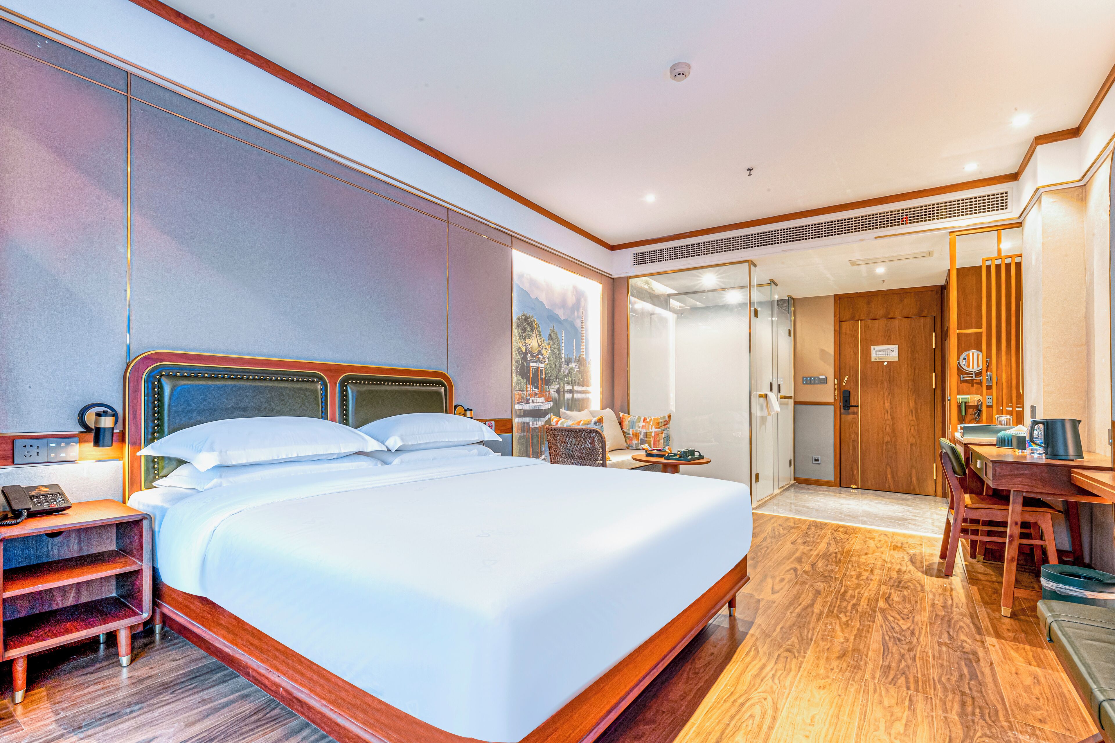 A hotel room with a large bed, a desk, and a chair. There is a small table with a lamp and a telephone.