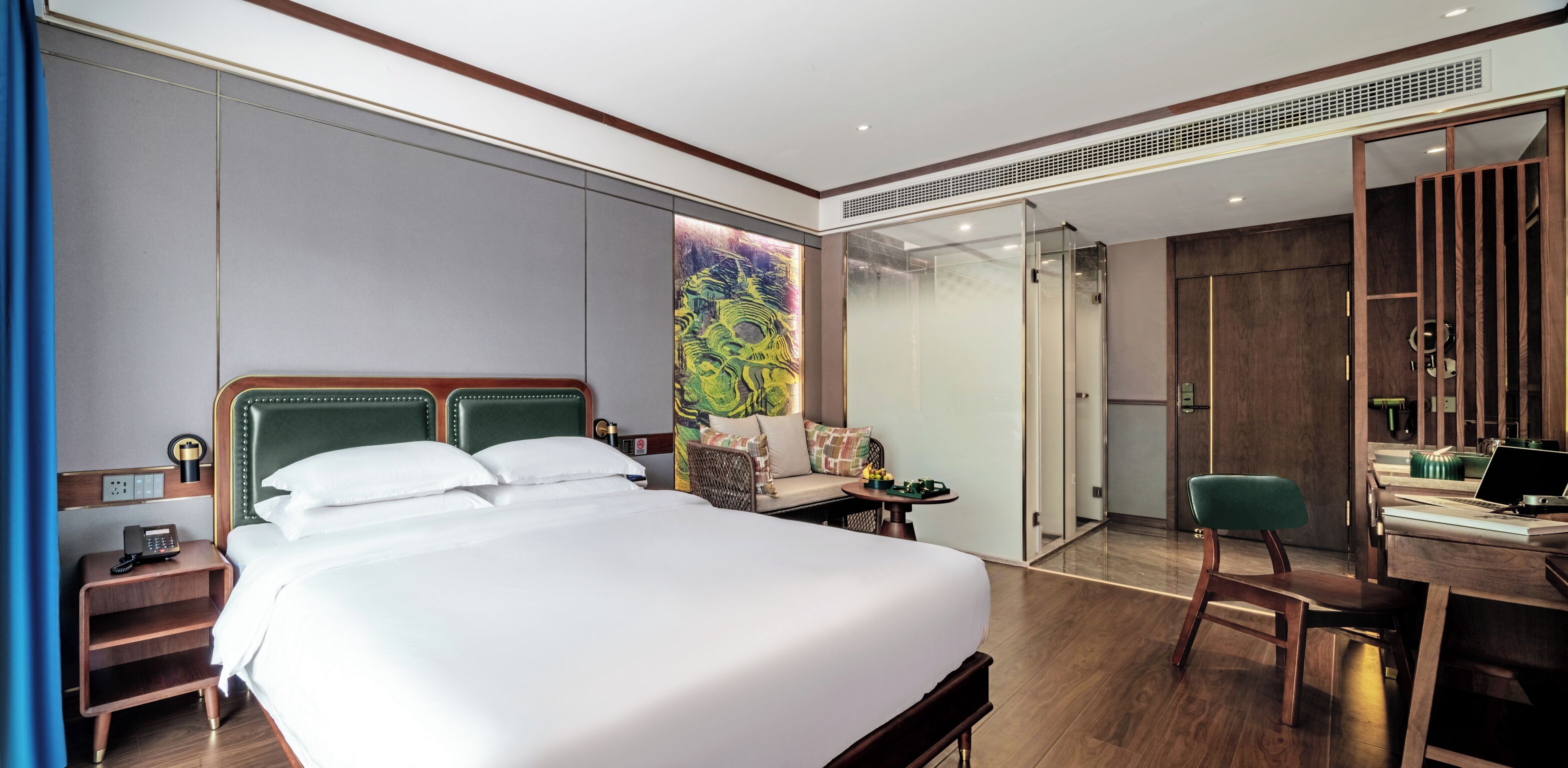 A hotel room with a large bed, a desk with a laptop, a chair, a small table with a lamp, and a sliding door leading to a bathroom.