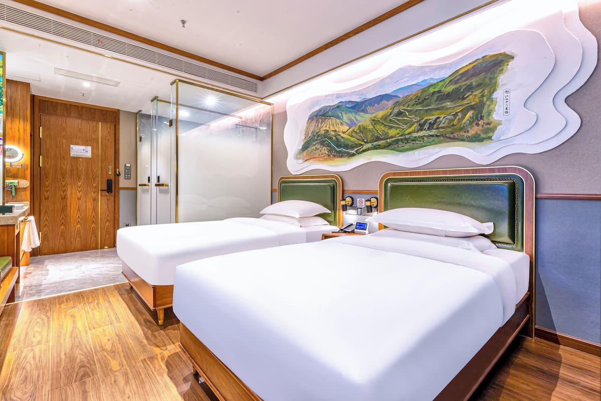 Une chambre d’hôtel avec deux lits, un plancher en bois, un tableau représentant un paysage accroché au mur et une cloison en verre qui sépare la chambre du couloir.