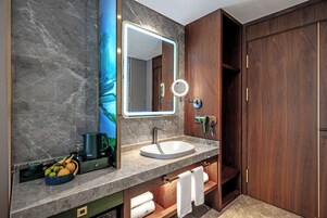 A modern bathroom with a marble countertop, a round mirror with a light, a sink, a coffee maker, and a wooden cabinet with towels.